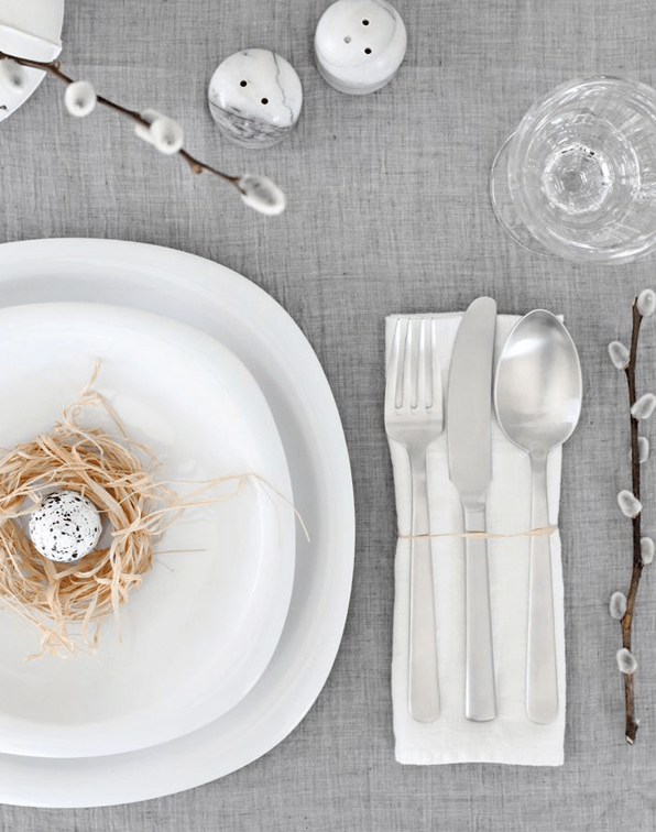 Tavola apparecchiata con tovaglia in lino grigia, gattici, uova di quaglia e nidi di paglia.