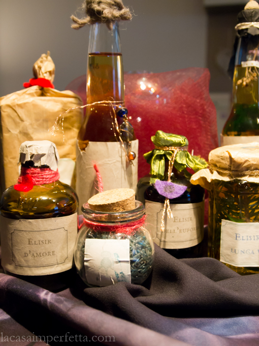 Primo piano di vasetti e bottiglie di vetro con pozioni varie, sullo sfondo la sfera di cristallo