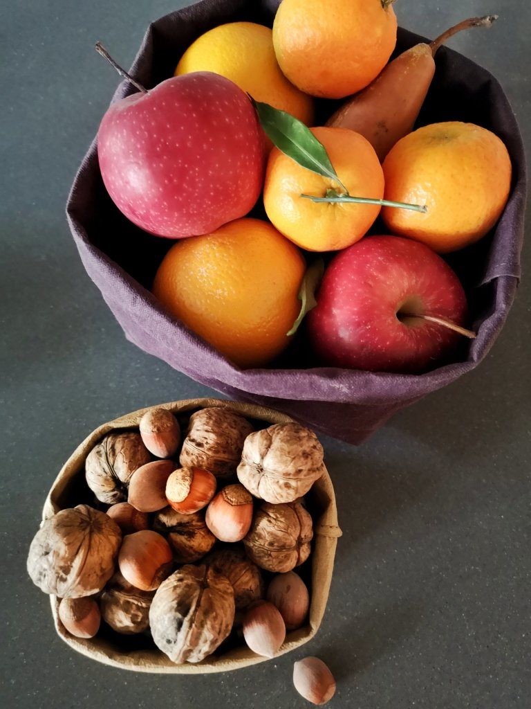 Due cestini porta frutta: uno viola con mele, mandarini e pere; uno piccolo color tabaccon con noci e nocciole nel guscio